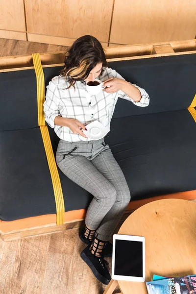 Vista de ángulo alto de la hermosa mujer joven sentada en el sofá y beber café - foto de stock