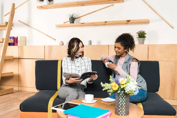 Happy young girlfriends drinking coffee and reading magazine in cafe — Stock Photo