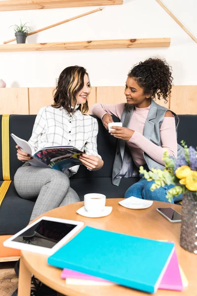 Stylish young girlfriends drinking coffee and reading magazine in cafe — Stock Photo