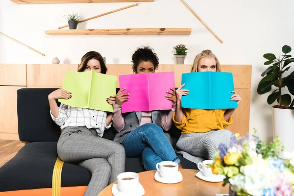 Group of young girlfriends covering faces with magazines in cafe — Stock Photo