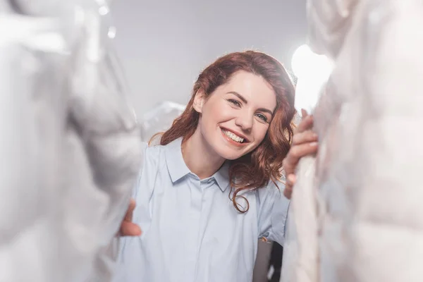 Lächelnde Reinigungskraft blickt in die Kamera zwischen Kleidungsstücken in Plastiktüten, die im Lager hängen — Stockfoto