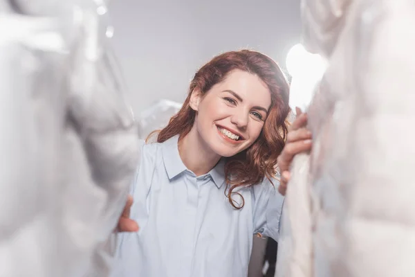 Travailleuse souriante de nettoyage à sec regardant la caméra entre les vêtements dans des sacs en plastique suspendus à l'entrepôt — Photo de stock