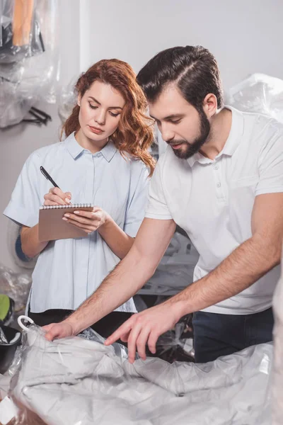 Serious dry cleaning workers with jacket in bag at warehouse — Stock Photo