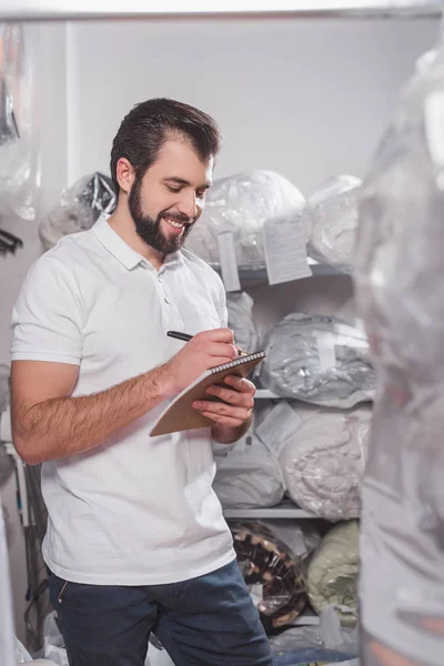 Souriant jeune travailleur de nettoyage à sec écrit dans le bloc-notes à l'entrepôt — Photo de stock