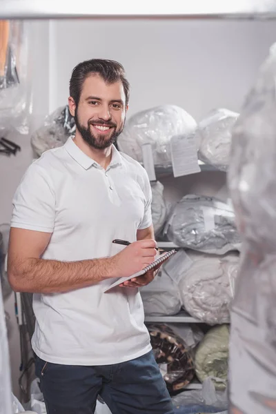 Apuesto trabajador de limpieza en seco escribir en bloc de notas en el almacén - foto de stock