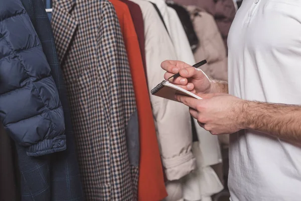 Cropped shot of dry cleaning worker with notepad counting clothes on hangers at warehouse — Stock Photo