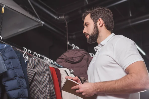 Travailleur de nettoyage à sec avec bloc-notes compter les vêtements sur les cintres à l'entrepôt — Photo de stock