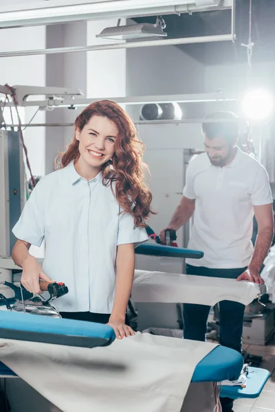 Happy young dry cleaning workers ironing clothes together — Stock Photo