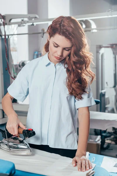 Female dry cleaning worker using industrial iron at warehouse — Stock Photo