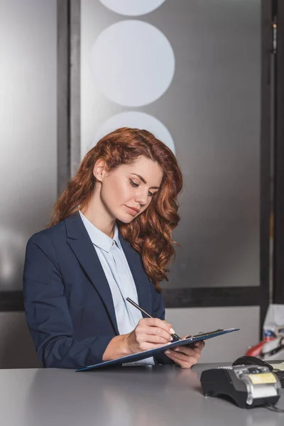 Young beautiful manageress writing in clipboard at workplace with pos terminal — Stock Photo