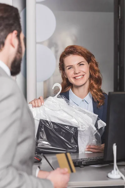 Dry cleaning manageress standing at workplace with bag of clothes while customer holding gold credit card — Stock Photo