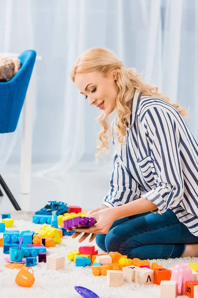 Vue latérale de la jeune mère avec des jouets assis sur le sol à la maison — Photo de stock