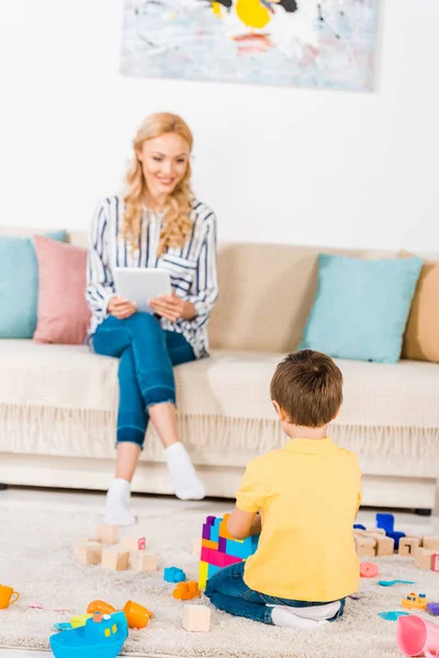 Маленький мальчик играет с игрушками в то время как мать использует планшет на диване дома — стоковое фото