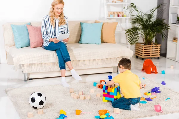 Menino brincando com brinquedos enquanto a mãe usando tablet no sofá em casa — Fotografia de Stock