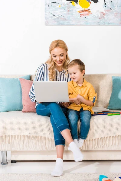 Madre sorridente e piccolo figlio utilizzando il computer portatile insieme sul divano di casa — Foto stock