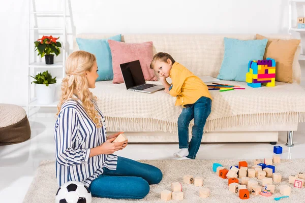 Menino usando laptop no sofá, enquanto a mãe sentado no chão em casa — Fotografia de Stock