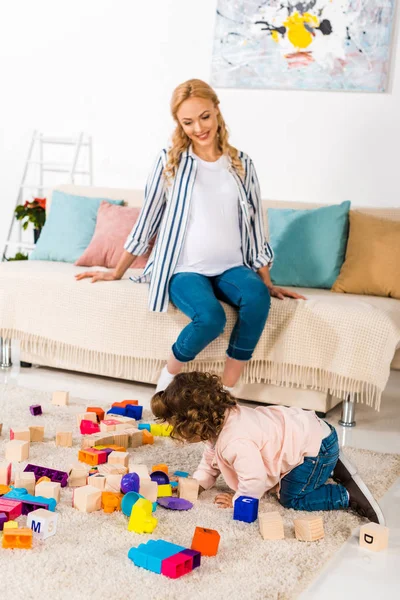 Sonriente embarazada madre buscando como hija jugando con juguetes - foto de stock