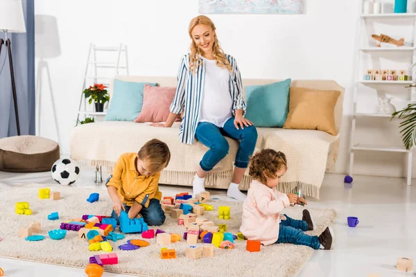 Heureuse mère enceinte regardant comment les enfants jouent avec des jouets — Photo de stock