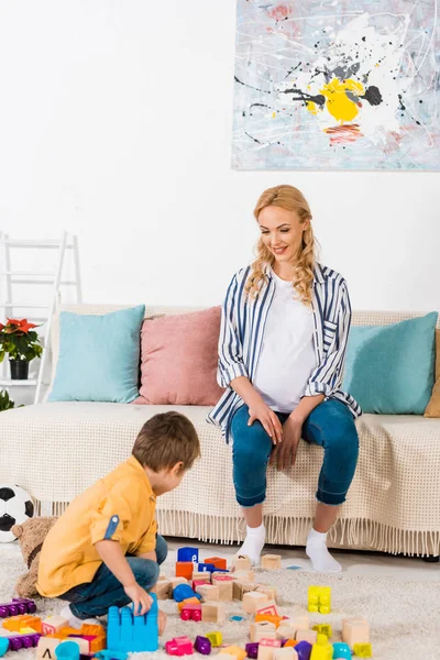 Feliz madre embarazada mirando cómo hijo jugando con juguetes - foto de stock