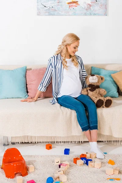 Smiling pregnant woman sitting on sofa with teddy bear — Stock Photo