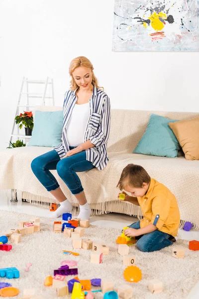 Happy pregnant mother looking how son playing plastic blocks — Stock Photo