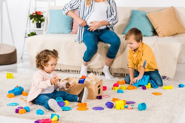 Обрезанный снимок беременной матери, сидящей на диване, и маленьких детей, играющих с красочными блоками дома — стоковое фото