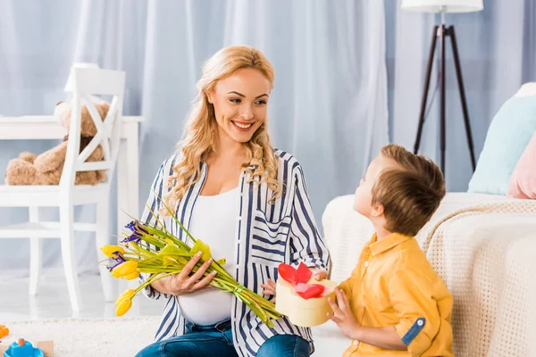 Adorable little boy presenting to happy pregnant mother tulip flowers and heart shaped gift box — Stock Photo