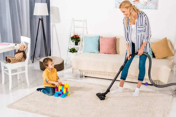 Mignon petit garçon jouant avec des blocs colorés et regardant mère enceinte nettoyage tapis avec aspirateur — Photo de stock