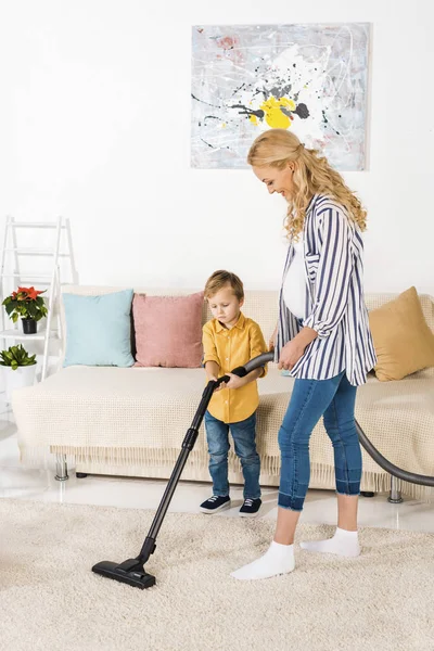 Smiling pregnant woman with adorable little son cleaning carpet with vacuum cleaner together — Stock Photo