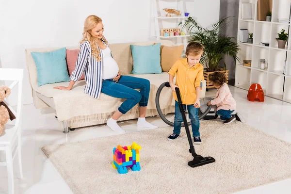 Mujer embarazada feliz mirando a los niños adorables habitación de limpieza con aspiradora en casa - foto de stock