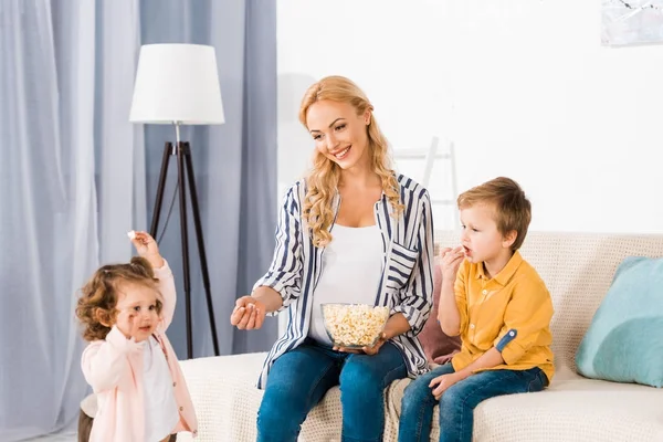 Happy pregnant mother and cute little kids eating popcorn from glass bowl at home — Stock Photo