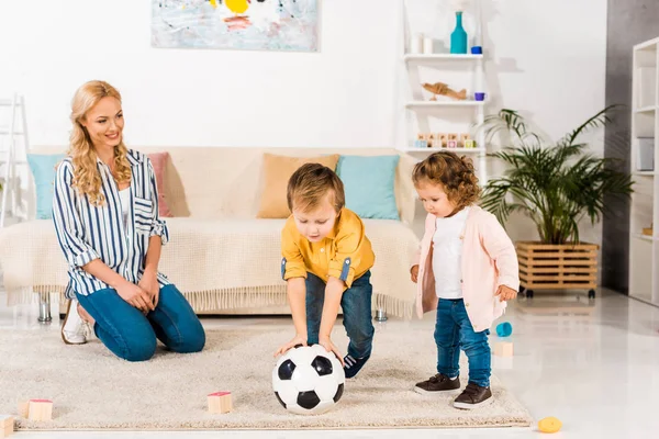 Sonriente madre mirando lindos niños pequeños jugando con pelota de fútbol en casa - foto de stock