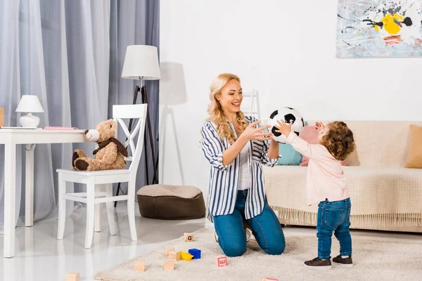 Feliz madre y linda hijita jugando con pelota de fútbol en casa - foto de stock