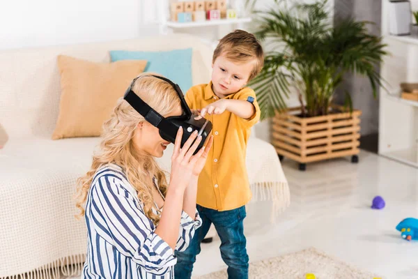 Adorable niño ayudando a la madre con auriculares de realidad virtual en casa - foto de stock