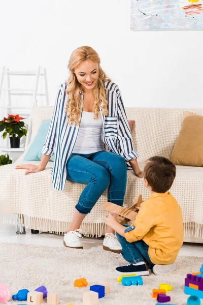 Mãe feliz olhando para o filho pequeno bonito brincando com avião de brinquedo em casa — Fotografia de Stock