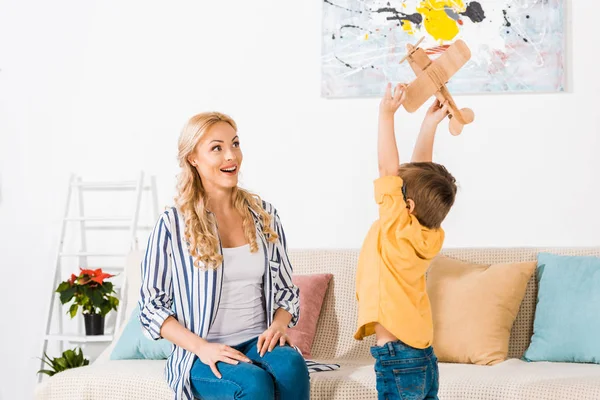 Cheerful mother looking at cute little son playing with toy plane at home — Stock Photo