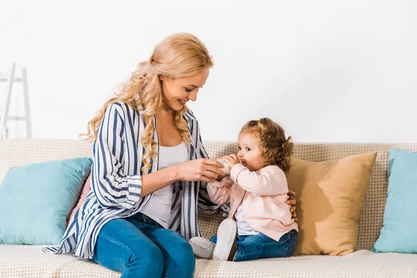 Mère heureuse nourrissant adorable petite fille avec du lait en bouteille à la maison — Photo de stock