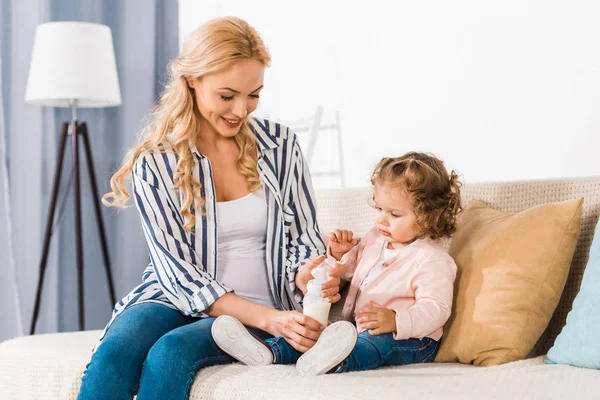 Schöne lächelnde Mutter füttert kleine Tochter zu Hause mit Milch in der Flasche — Stockfoto