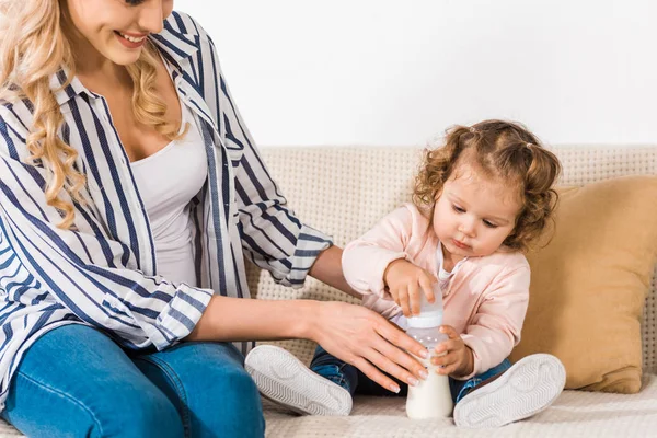 Schnappschuss einer lächelnden Mutter, die auf dem Sofa sitzend ihre kleine Tochter mit der Flasche anschaut — Stockfoto