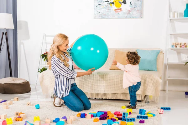 Sonriente madre y adorable hijita jugando con bola en forma y bloques de colores en casa - foto de stock