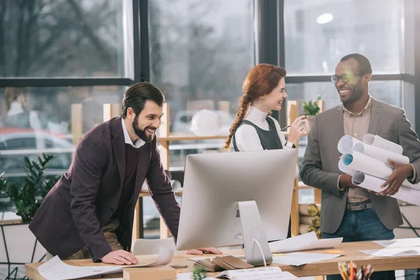 Arquitectos multiétnicos sonrientes que trabajan con planos y computadoras en la oficina moderna - foto de stock