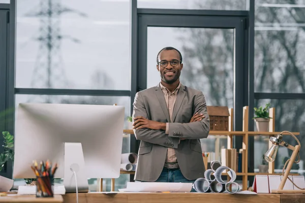 Sorridente arquiteto afro-americano com braços cruzados em pé no local de trabalho com computador e plantas — Fotografia de Stock