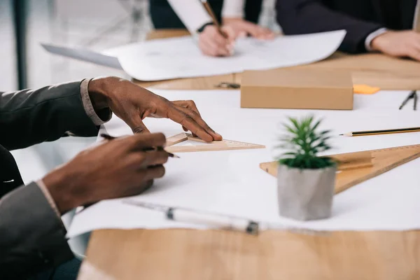 Cropped view of multiethnic architects drawing blueprints with rulers in office — Stock Photo