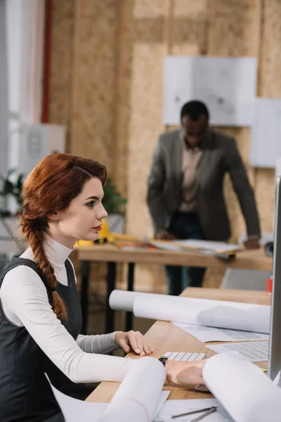 Hermosa mujer arquitecta utilizando la computadora mientras su colega de pie borrosa en el fondo - foto de stock