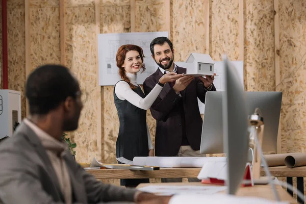 Junge Architekten zeigen ihrem Kollegen das Modell eines Hauses, während er mit dem Computer arbeitet — Stockfoto
