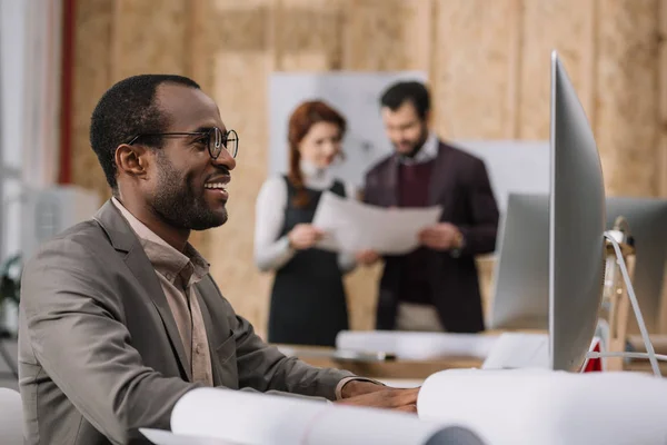 Щасливий афроамериканський архітектор, який працює з комп'ютером в сучасному офісі, а його колеги обговорюють контракт — стокове фото