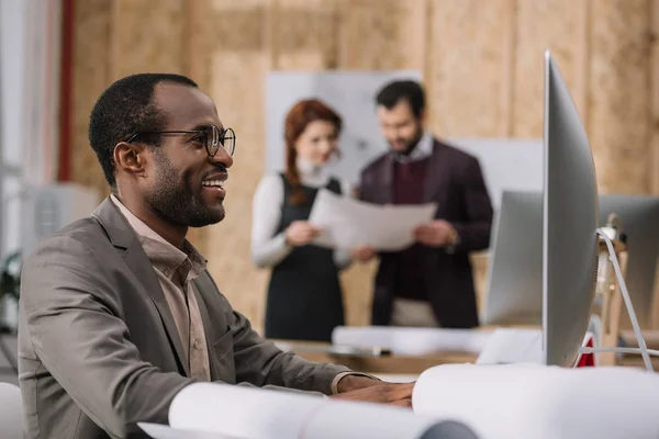 Felice architetto afroamericano che lavora con il computer in ufficio moderno mentre i suoi colleghi discutono di contratto — Foto stock