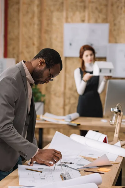 Konzentrierter afrikanisch-amerikanischer Architekt zeichnet Baupläne, während sein Kollege das Modell eines Papierhauses entdeckt — Stockfoto