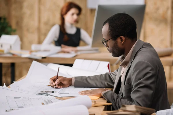 Apuesto arquitecto afroamericano dibujo planos del edificio mientras que su colega que trabaja con la computadora - foto de stock