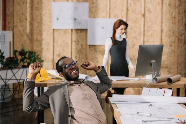 Handscome arquiteto afro-americano que se estende na cadeira enquanto seu colega de trabalho com computador — Fotografia de Stock